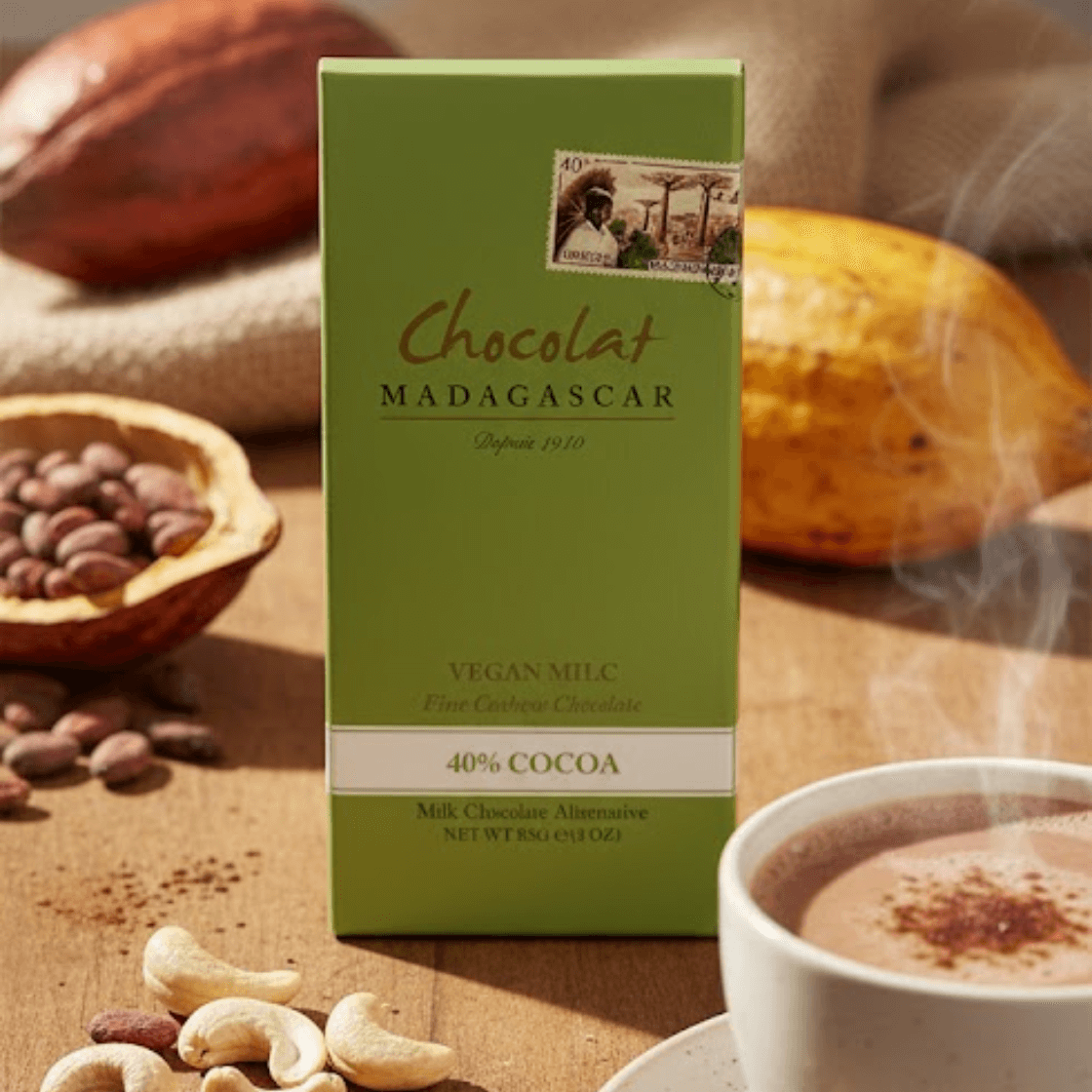 Teplá, vertikálna životná snímka vysokej, zelenej krabice Chocolat Madagascar Vegan MILC 40% Cocoa Fine Cashew Chocolate stojacej vzpriamene na rustikálnom drevenom povrchu. Roztrúsené kešu ležia v popredí vedľa parného šálky horúcej čokolády na malom tanieriku. V pozadí sú surové kakaové bôby v drevenej miske a celé kakaové struky odpočívajúce na jutovej látke, osvetlené jemným slnečným svetlom.